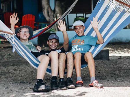 Three students sitting in a hammock smiling and waving
