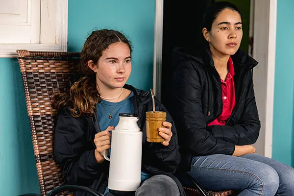 Volunteer holding a thermos and cup of tereré while sitting next to a host family member